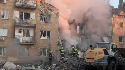 Dmitry Vlasov Three people in fluorescent clothing and helmets climb a mountain of rubble, the remnants of a residential building that has collapsed. Smoke is billowing from the rubble, tinted pink by the early morning sunrise.