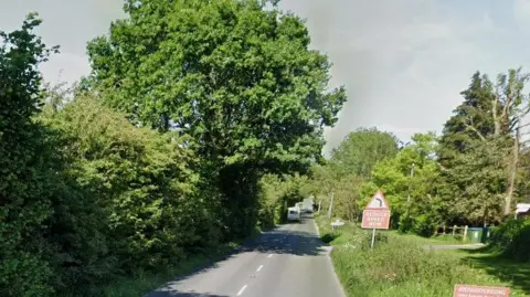 Google A Google Street view image which shows a tree-lined street with one lane in each direction. There is a reduce speed now sign on the verge on the right side of the road.