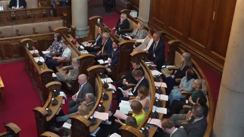 Liberal Democrat councillors sit on the curved wooden benches inside Hull's Guildhall council chamber. Leader Mike Ross is on his feet talking