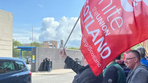 People protesting outside a Stagecoach depot with a Unite union red banner. There is a photographer taking pictures across the street 