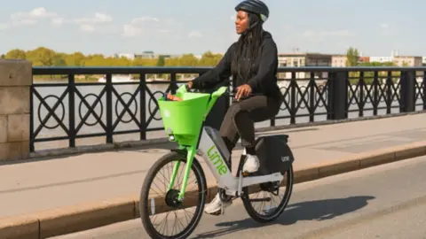 A woman rides a bike with green Lime insignia on it. There is a green basket on the bike as well. She is wearing a black helmet and black clothing and is cycling on a flat road near a river. 