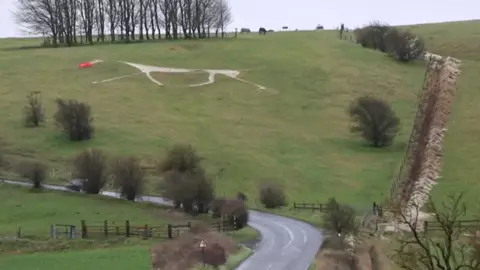 A thin white horse on a hillside with red fabric on its note. A road meanders in front of it.