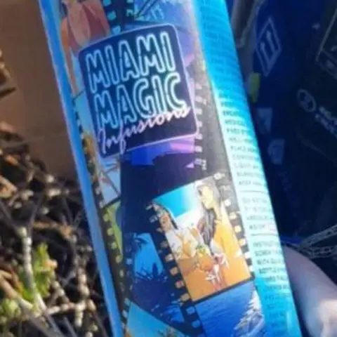 Welsh Cottage by Zina A close-up photo of a circular blue tin of nitrous oxide being held in someone's right hand. The tin is decorated with filmstrip images and has the label "Miami Magic Infusions'