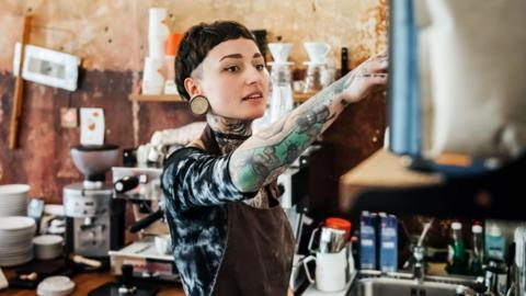 A woman with tattooed arms and neck working in a coffee shop