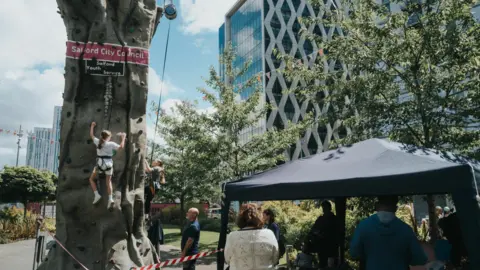 We Invented The Weekend Large oblong climbing wall with a couple of children scaling it. The wall is outdoors in Media City.