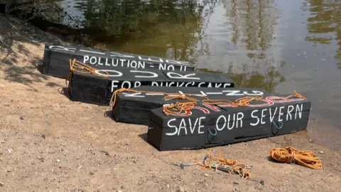 Three coffins sit on the banks of the river. They are decorated with the words Save Our Severn.