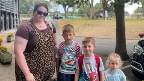 Alana - a woman on the left of the photo, who has brown hair and blue sunglasses, wearing a leopard print overall and purple red T-shirt. Next to her is her son, with lighter blonde hair wearing a chequered red backpack and grey race car t shirt. Next to him is her other son, wearing a grey Disney T-shirt and a red backpack. Next to him is her daughter with blonde hair wearing a blue and white t shirt. In the background is a car park and field.