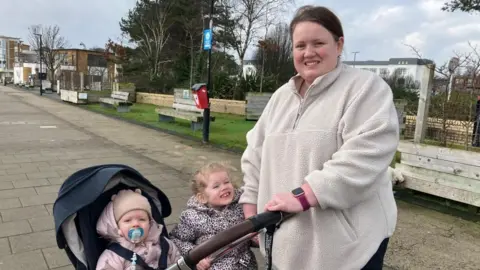 A woman is standing with two children. One is in a buggy and is wearing a light-coloured wooly hat. There are wooden benches in the background.