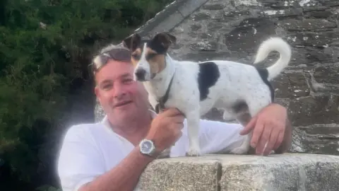James Southey looking and smiling at the camera with his small dog stood on a wall.