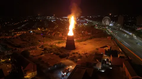 Flames are coming out of a tower, silhouetted against a dark sky. You can see some rooftops and street lights.