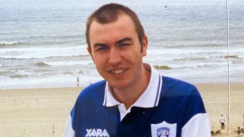 Val White Martin has short brown hair and is wearing a blue and white Cardiff City shirt. Behind him is the beach and shoreline.