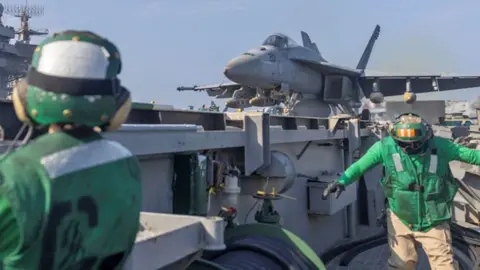 @USAfricaCommand Two men in bright green jackets and helmets can been see in with a US army jet behind them - image released by US Africa Command, February 2025