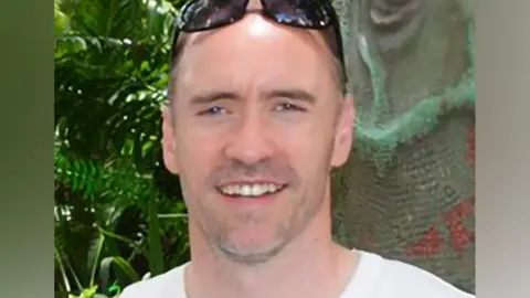 Paul Doyle has short dark greying hair with sunglasses on his head and is wearing a white T-shirt.