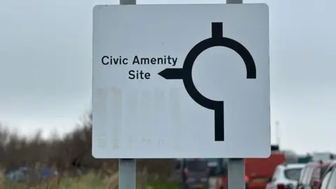 A wite and black road sign with an arrow to the site. Al row of cars waits in a queue alongside it.
