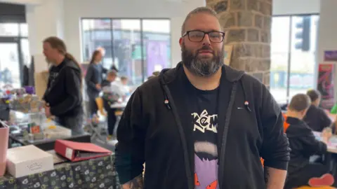 A man with a black and grey beard, short grey hair and black-rimmed glasses is wearing a black hoodie over a Pokemon t-shirt. In the background people are playing table-top games.