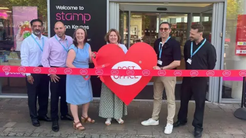 Post Office A group of people, standing outside a shop, holding a large red heart, with a red ribbon in front of them. They are all standing. A woman in a blue dress is holding a large pair of red scissors. 