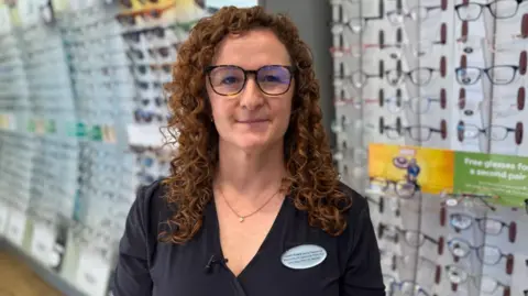 Lowri Evans, in a black dress, with glasses on, looks at the camera. It is a head and shoulders shot of her.