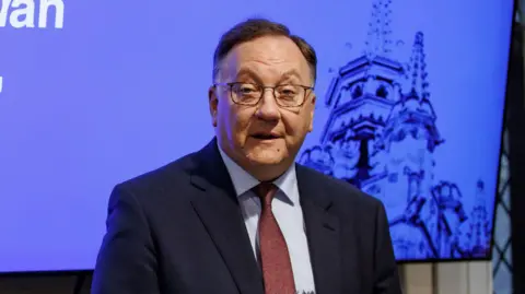 PA Media A man, dressed in a dark suit, blue shirt and red tie, is speaking in front of a television screen. It has a blue background and a dark image showing the steeple of a church. The man has short, dark hair which is swept to the side and is wearing glasses. 