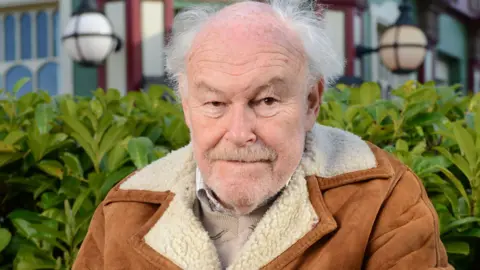 Actor Timothy West looks at the camera. He has a grey beard and grey hair. He is wearing a tan coloured coat with a sheepskin collar.