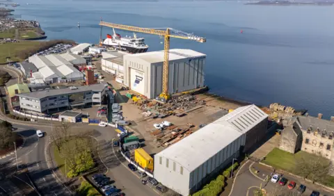 An aerial view of a shipyard beside a wide river. Several light grey fabrication facilities can be seen and a large yellow crane and a large ferry is moored at the quayside in the background