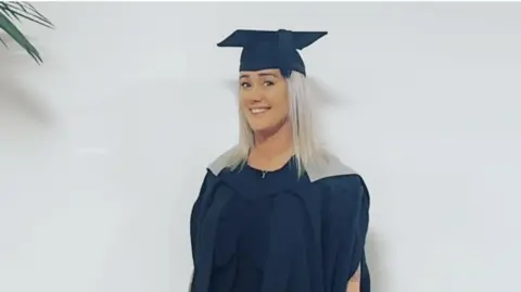 Keziah Hall A woman with blond hair posing in a university graduation hat and cape.