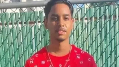 Shawn Seesahai wearing a red t-shirt and neck chain. He is standing in front of a green fence.