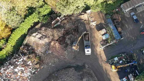 An aerial shot of lorries and waste on a brown patch of land surrounded by trees