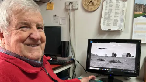 A man with a grey looks at the camera smiling while sitting in front of a computer screen with historic images of planes.