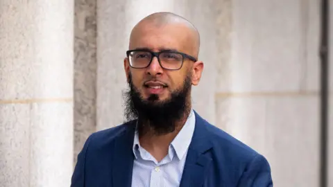 Majid Novsarka wearing a blue suit and a blue and white shirt as well as dak rimmed glasses 