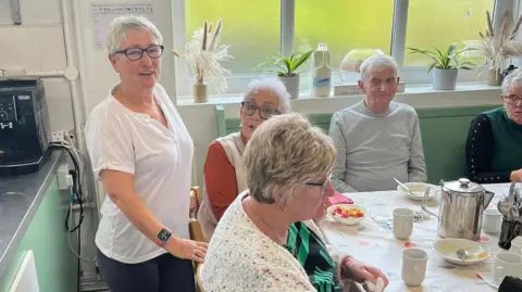 Cortonwood Comeback A woman with short white hair, black glasses and a white polo shirt smiles at the camera. She is standing behind a number of older people sat around a table. 