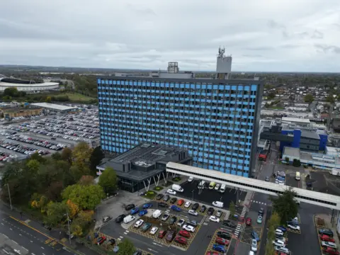 An aerial view of a large hospital tower block featuring blue panels, grey cladding and windows. The building is surrounded by car parks, vehicles and nearby roads. A football stadium is visible in the distance.