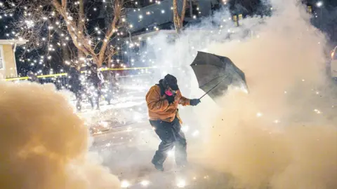 A person wearing a mask and other protective clothing holds up an umbrella in a smoky street after federal agents fire munitions and pepper balls in north Minneapolis.