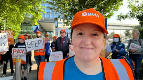 Amanda White/BBC A woman with her hair tied back wearing a orange BMA hat and orange hi-vis top with a blue top underneath. She is smiling at the camera. Behind her are several people holding up signs.
