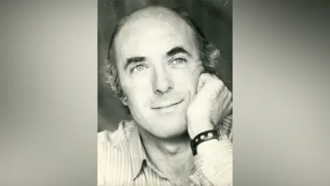 Daniel Hicks A black and white image of a man who rests his head in hand. He wears a shirt and there is a small watch around his wrist. 