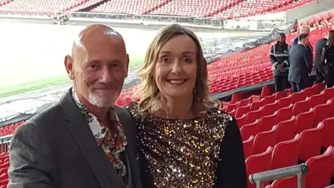 Ruth and Peter Doyle smiling at the camera in the seating of an almost empty sports stadium. Ruth wears a sparkly dress and Peter has a jacket and floral shirt.