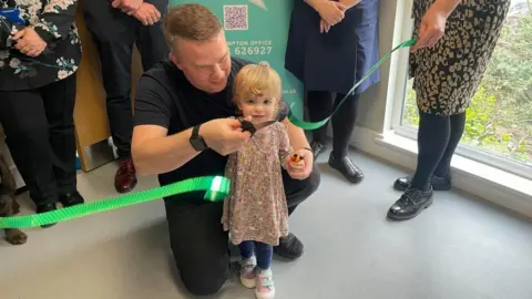Kate Bradbrook/BBC Chloe holds the scissors with her father kneeling beside her, having just cut a green ribbon as part of the hospital's unveiling