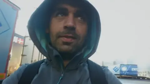 BBC/The Open University The same man as in the first picture, wearing a big coat and hood, walking a shipping yard. He looks tired.