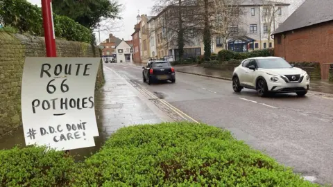 Route 66 Potholes sign alongside a town centre road with cars going by in each direction