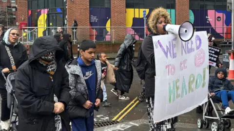 Barton House: Adults and children marching with a woman at the front talking into a megaphone and holding a sign that says 'We need to be heard'