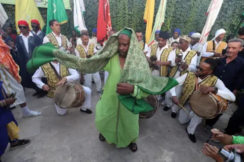 EPA A man dressed in a traditional green outfit dancing in the middle of a circle of men who are also wearing tradition white and yellow outfits and playing drums.