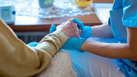 Getty Images/Sanja Radin Health worker holding hand of elderly woman