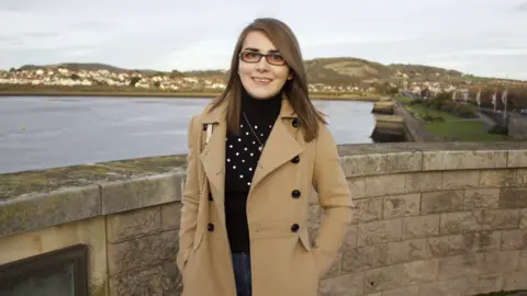 Williams family Elin standing in front of a wall with the sea behind her and houses and hills further away in the distance