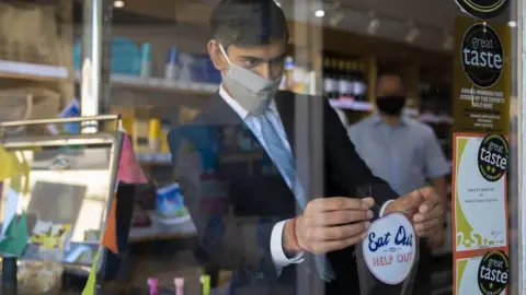 HM Treasury Then-Chancellor Rishi Sunak applies a "Eat Out to Help Out" sticker to a window of a business in Northallerton