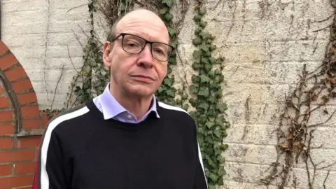 Alastair France standing in front of a wall at his home