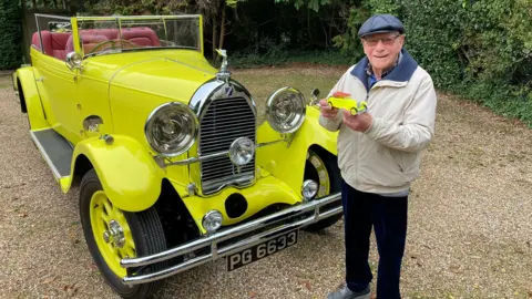 Justin Dealey/BBC Malcolm Stern standing in front of a classic car, holding a 3D printed version of the car