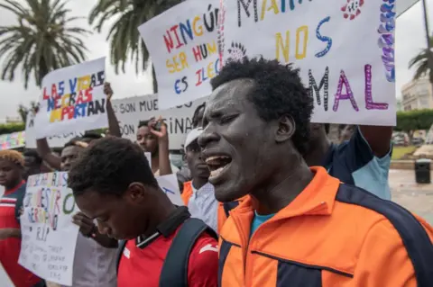Getty Images Protesters hold placards during the demonstration. Dozens of people gathered at the Plaza de España in the city of Melilla to demand the action of the security forces of both countries in the massive entry attempt by hundreds of migrants, mainly from sub-Saharan Africa. The demonstration was also joined by Spanish citizens, members of civil society, and some humanitarian organizations. Today marks a week of the events where 23 people lost their lives, dozens ended up with injuries, and the Moroccan police detained many others at the border crossing known as Chinatown within Moroccan territory.
