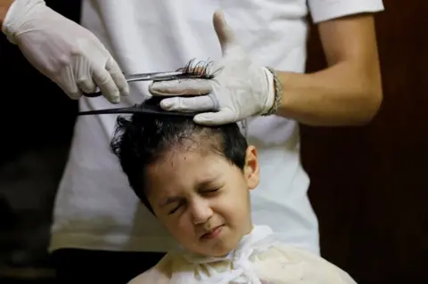 Reuters A boy grimaces as his hair is clasped and trimmed by a gloved barber.