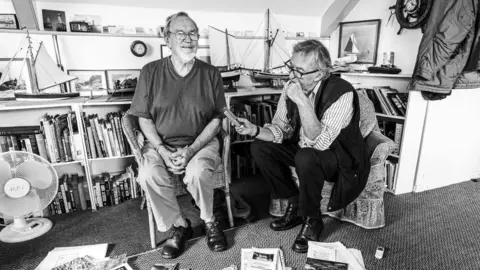 Andrew Hobbs Photography Black and white image of Tony James interviewing someone. They are sat side by side on chairs. Shelves crammed full of books can be seen behind them, as well as on the floor.