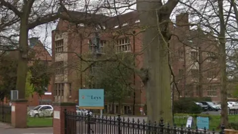 Google An exterior shot of The Leys school in Cambridge taken from the road. It shows a large brick building in the background with trees in the foreground and a blue school sign.