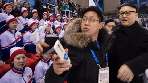 Getty Images Some cheerleaders look at a Kim Jong-un impersonator disapprovingly whilst others look at the hockey match